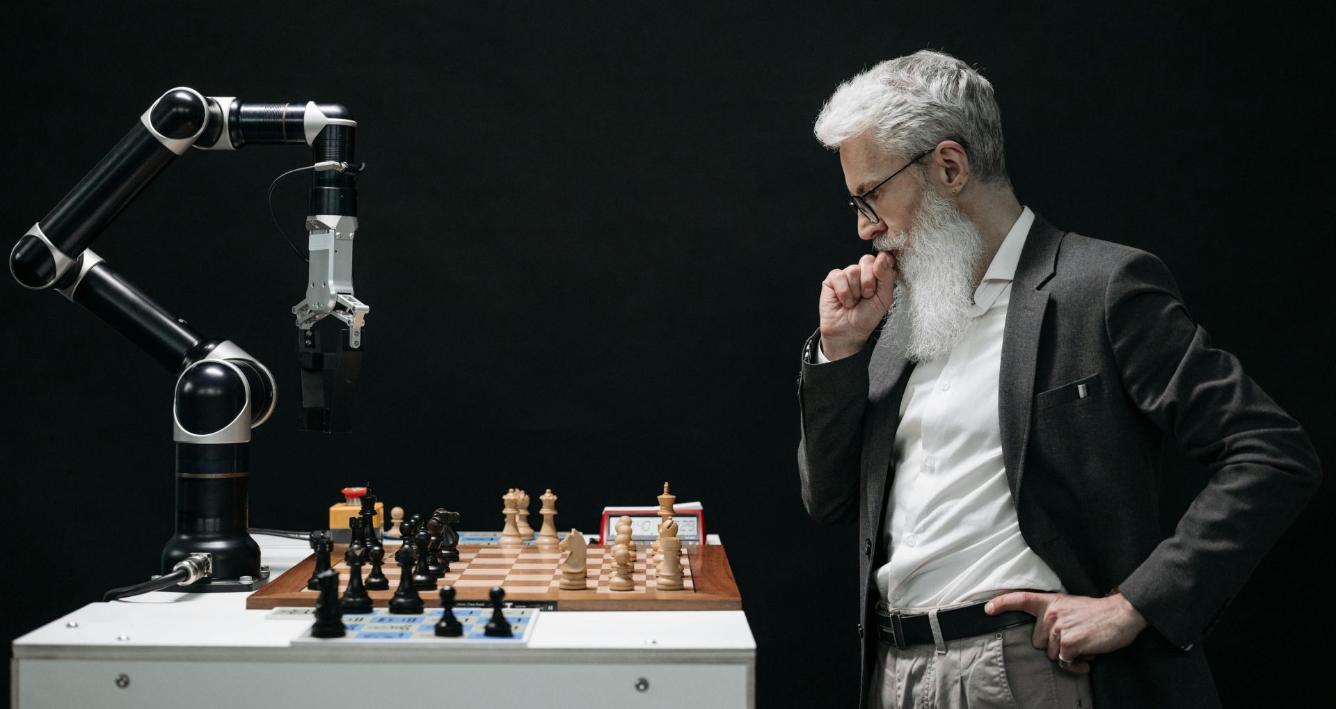 An elderly scientist contemplates a chess move against a robotic arm on a chessboard.